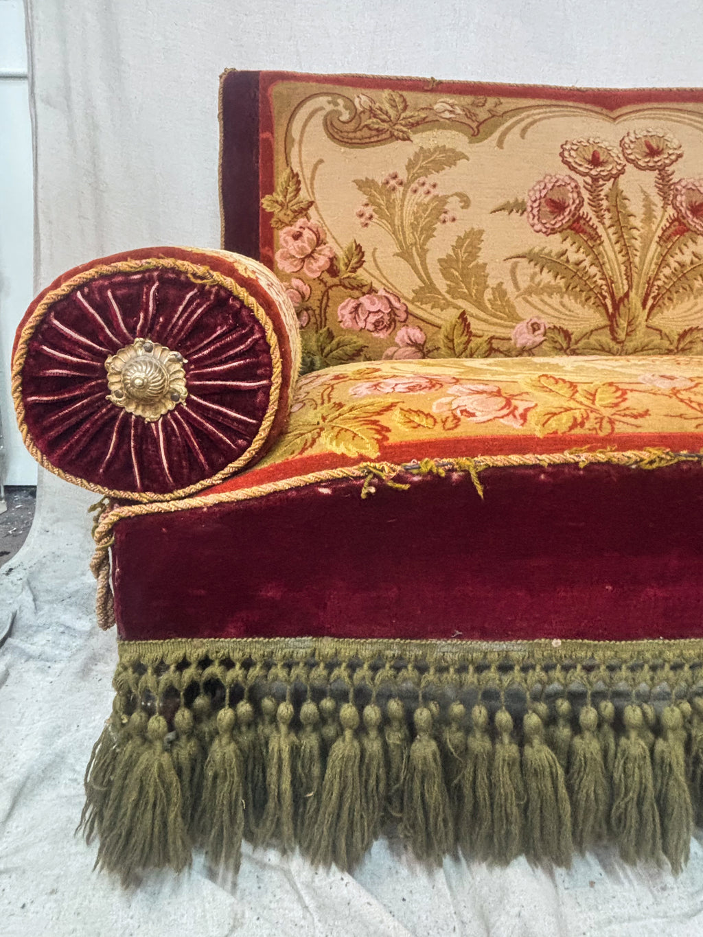 An antique Victorian-style loveseat with red velvet trim, a beige floral tapestry back and seat, two red bolster pillows, and olive green fringe along the bottom.