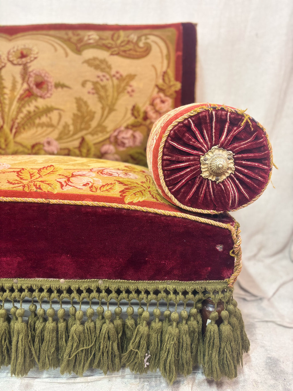 An antique Victorian-style loveseat with red velvet trim, a beige floral tapestry back and seat, two red bolster pillows, and olive green fringe along the bottom.