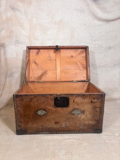 An antique wooden trunk with an open lid, showing a warm brown wood grain interior and exterior, decorated with brass corner brackets and a central latch, set against a draped beige fabric backdrop.