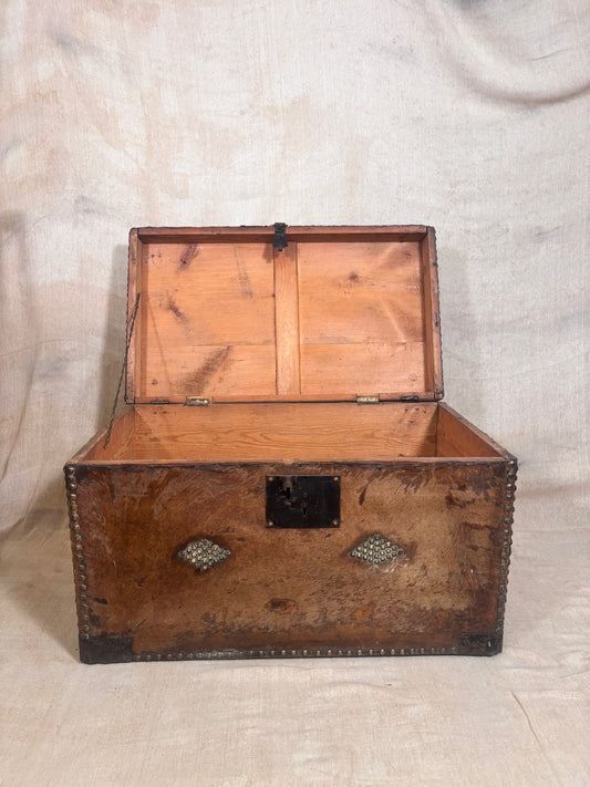 An antique wooden trunk with an open lid, showing a warm brown wood grain interior and exterior, decorated with brass corner brackets and a central latch, set against a draped beige fabric backdrop.