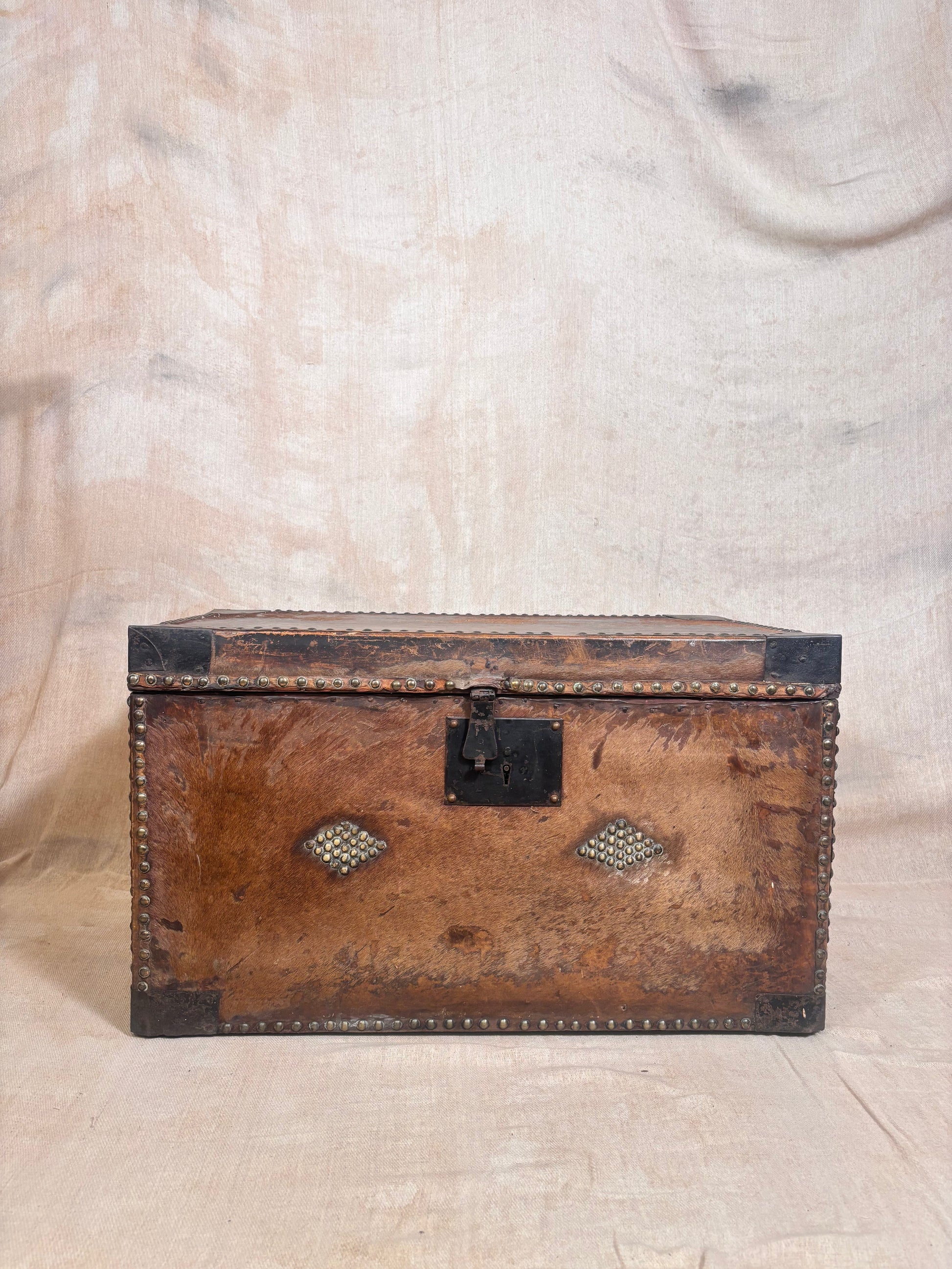 An antique brown leather trunk with black metal corner protectors and a central latch, decorated with two diamond-shaped metal medallions on the front, set against a draped beige fabric backdrop.