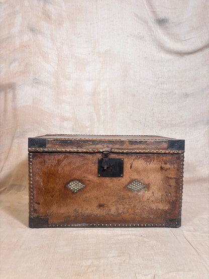 An antique brown leather trunk with black metal corner protectors and a central latch, decorated with two diamond-shaped metal medallions on the front, set against a draped beige fabric backdrop.