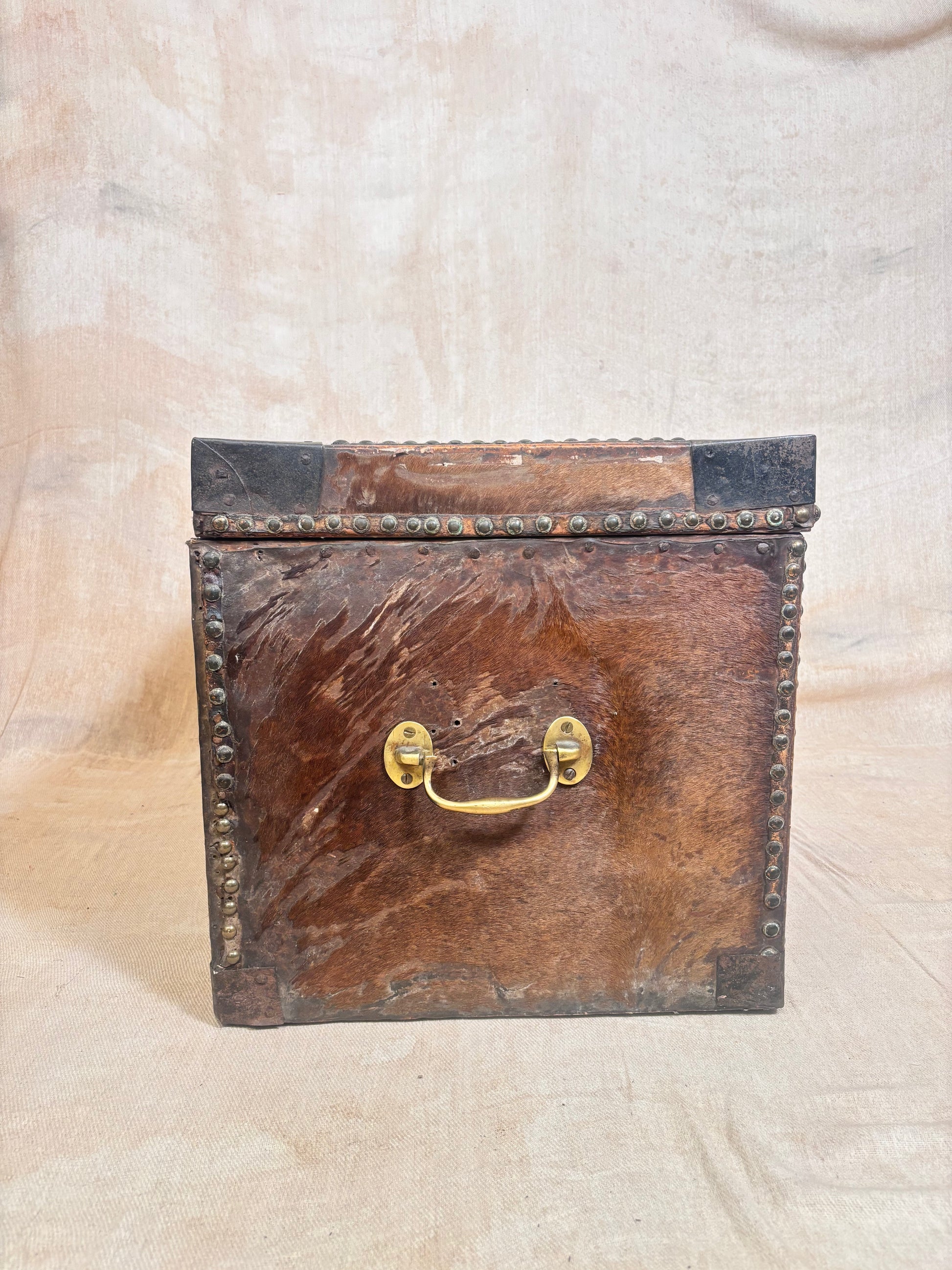 A vintage rectangular leather trunk or storage box with a distressed brown leather front, dark corner protectors, brass hardware handles, and metal studs along the edges, set against a neutral beige fabric backdrop.