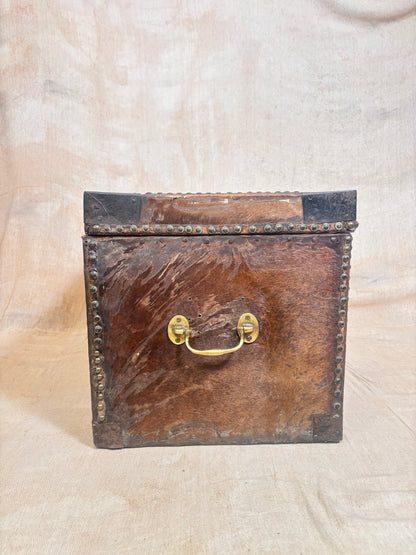 A vintage rectangular leather trunk or storage box with a distressed brown leather front, dark corner protectors, brass hardware handles, and metal studs along the edges, set against a neutral beige fabric backdrop.