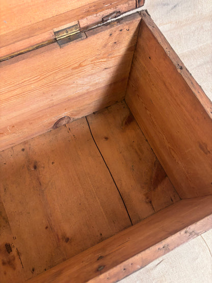 An open wooden storage chest or trunk made of warm medium-brown wood, showing the interior grain, a metal hinge, and the bottom corner against a light neutral floor.