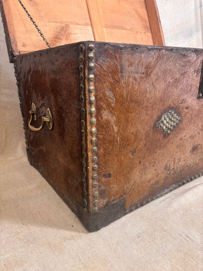 An antique brown leather trunk with brass hardware, nail trim, and a hinged lid, shown on a neutral fabric background.