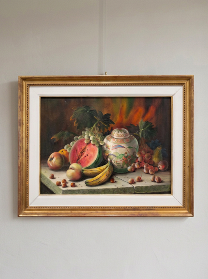 A framed still life painting of fruit and a ceramic jar, shown hanging on a light gray wall. The artwork features a gold frame with a white mat, and the painting depicts watermelon, grapes, bananas, apples, and a decorative vase.
