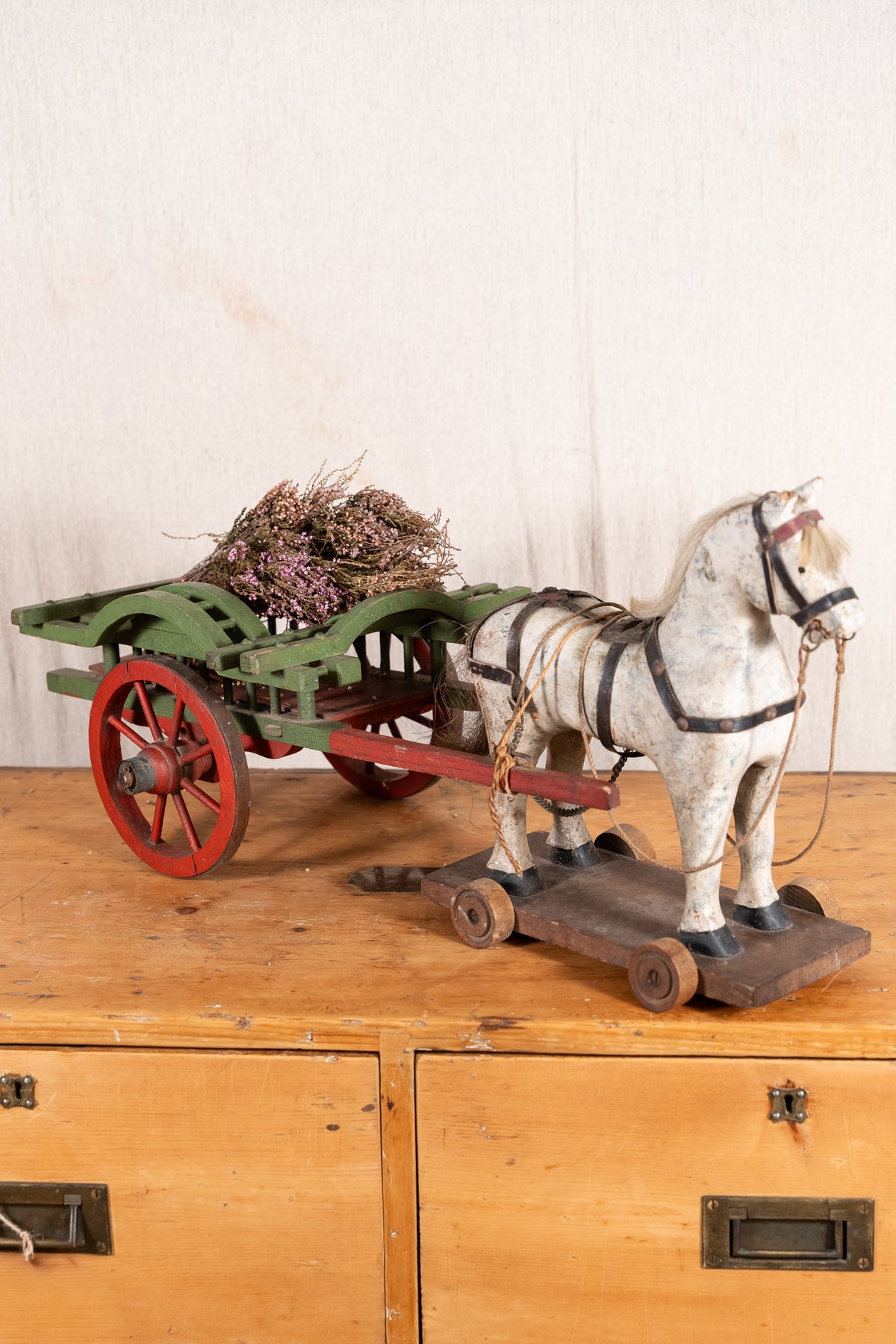 A vintage model of a white horse pulling a green cart with red wheels, filled with dried heather, displayed on a wooden cabinet against a light wall.