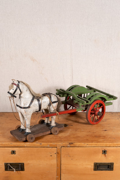 A vintage toy horse-drawn cart set on a wooden surface against a textured off-white wall. The toy features a white horse with black harness details on a wooden base, pulling a green cart with red wheels.