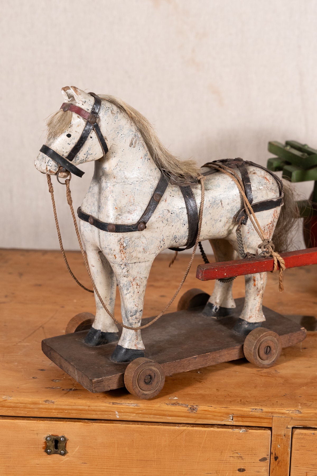 A vintage wooden toy horse on a wheeled cart, painted in distressed off-white with black harness details and a light brown mane, displayed on a wooden surface against a neutral background.