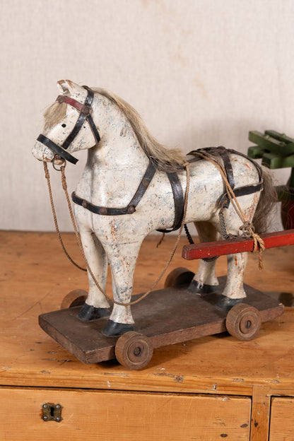 A vintage wooden toy horse on a wheeled cart, painted in distressed off-white with black harness details and a light brown mane, displayed on a wooden surface against a neutral background.