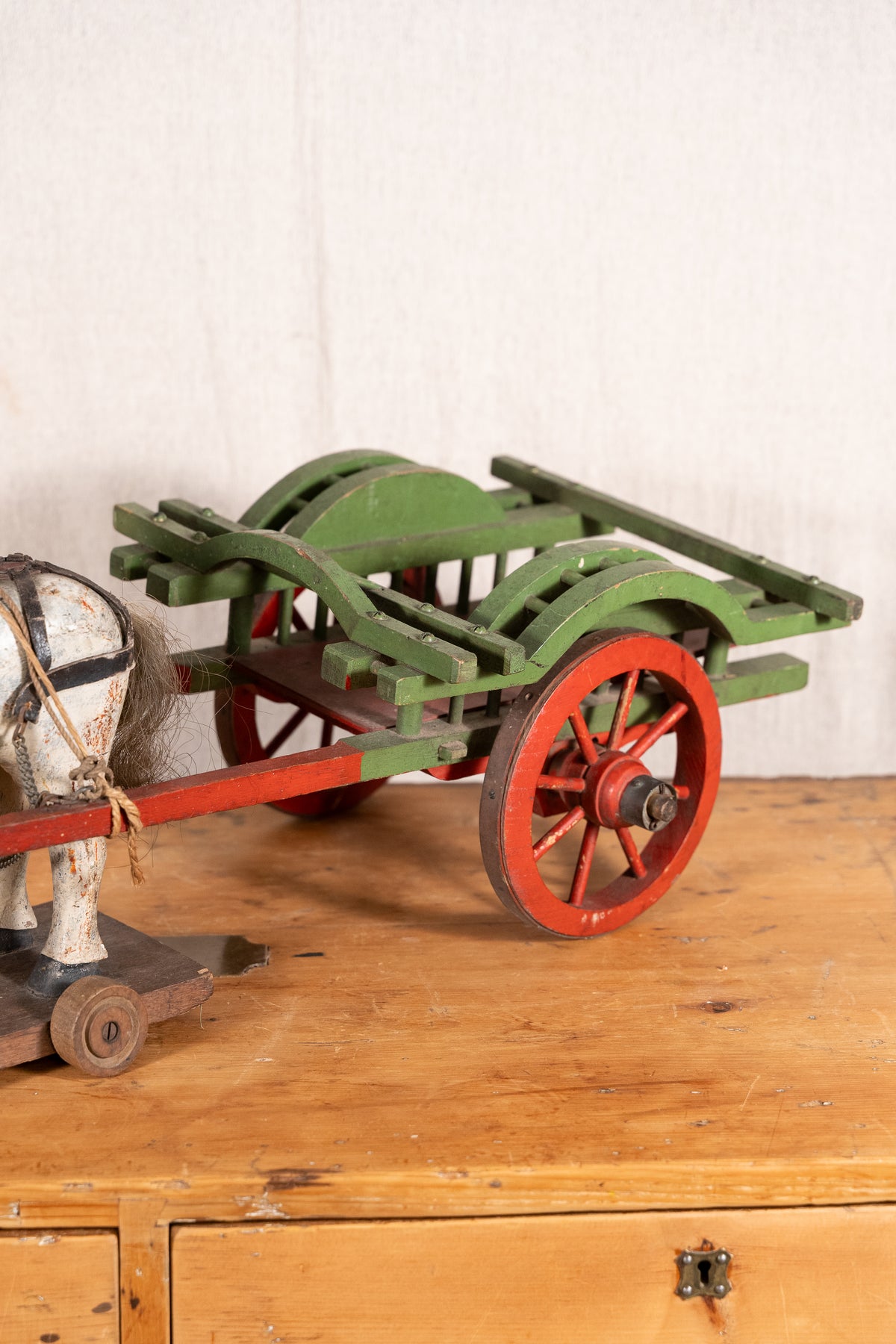 A vintage toy wooden cart with green body panels and red spoked wheels, shown on a wooden surface against a light wall. Part of a horse figure is visible at the left edge.