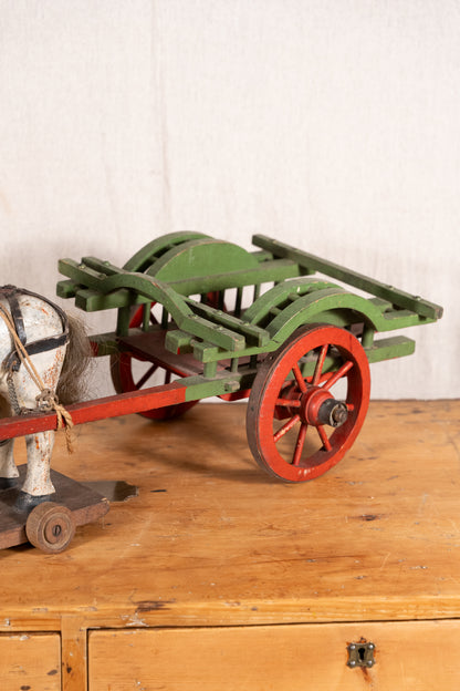 A vintage toy wooden cart with green body panels and red spoked wheels, shown on a wooden surface against a light wall. Part of a horse figure is visible at the left edge.