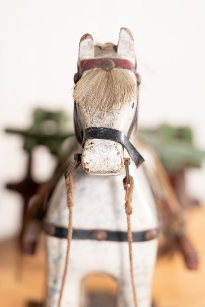 A vintage painted wooden toy horse with a distressed white finish, black bridle details, and light brown faux hair, shown in a close-up shot with a warm, blurred background.