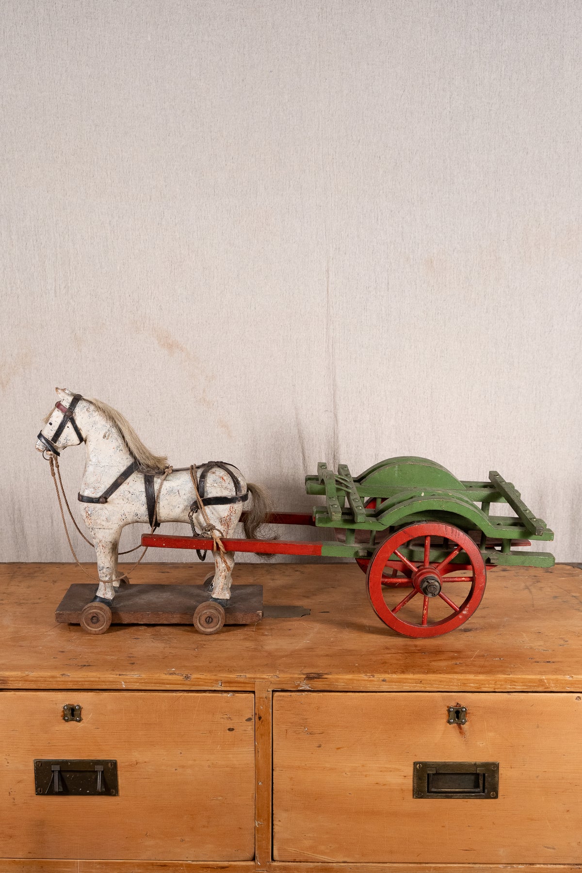 A vintage toy horse-drawn cart, featuring a white wooden horse with black harness details and a green cart with red wheels, displayed on a wooden chest against a textured off-white wall.