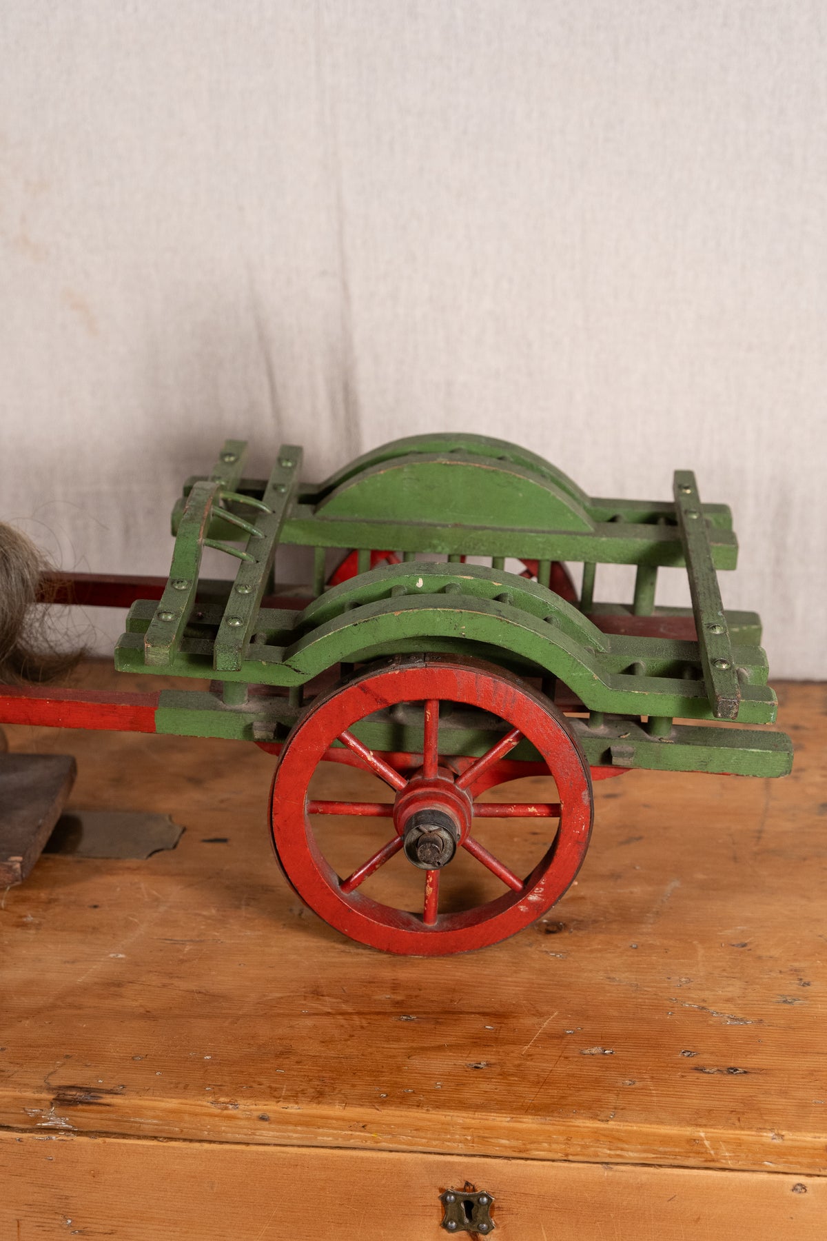 A vintage green wooden cart with red-painted wheels and a red handle, displayed on a rustic wooden table against a light fabric backdrop.