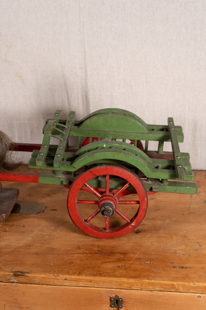 A vintage green wooden cart with red-painted wheels and a red handle, displayed on a rustic wooden table against a light fabric backdrop.