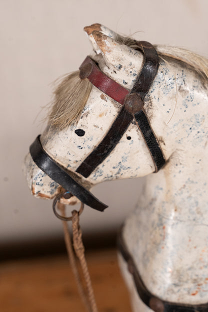 Close-up of a vintage wooden rocking horse head with a distressed off-white painted finish, black leather-like straps, and a reddish-brown bridle detail.