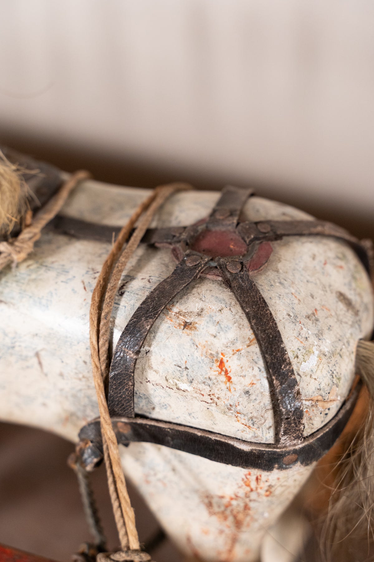 Close-up of a vintage white wooden toy horse with dark leather straps and rope tack, showing weathered details and a blurred neutral background.