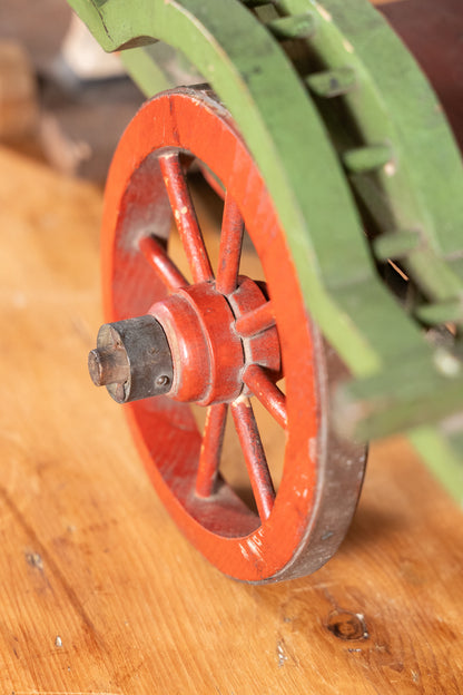 A close-up of a vintage red wooden spoked wheel with a dark metal hub, resting on a wooden surface with a green machine part blurred in the background.