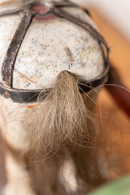 Close-up of a textured, distressed white hat or headpiece with dark straps and a long, light brown hair tuft hanging in the center, set against a warm, blurred background.