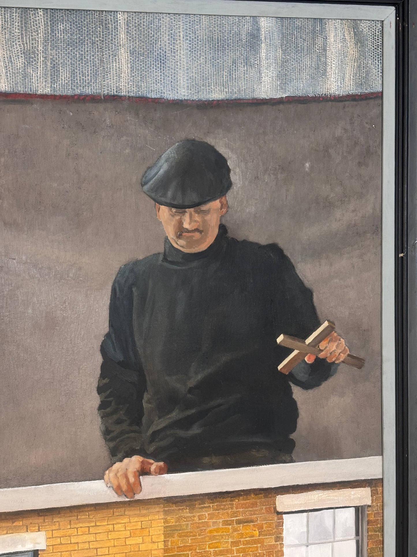 A framed painting depicting a man in a black cap and black turtleneck sweater holding a wooden cross, leaning over a brick wall under a gray background.