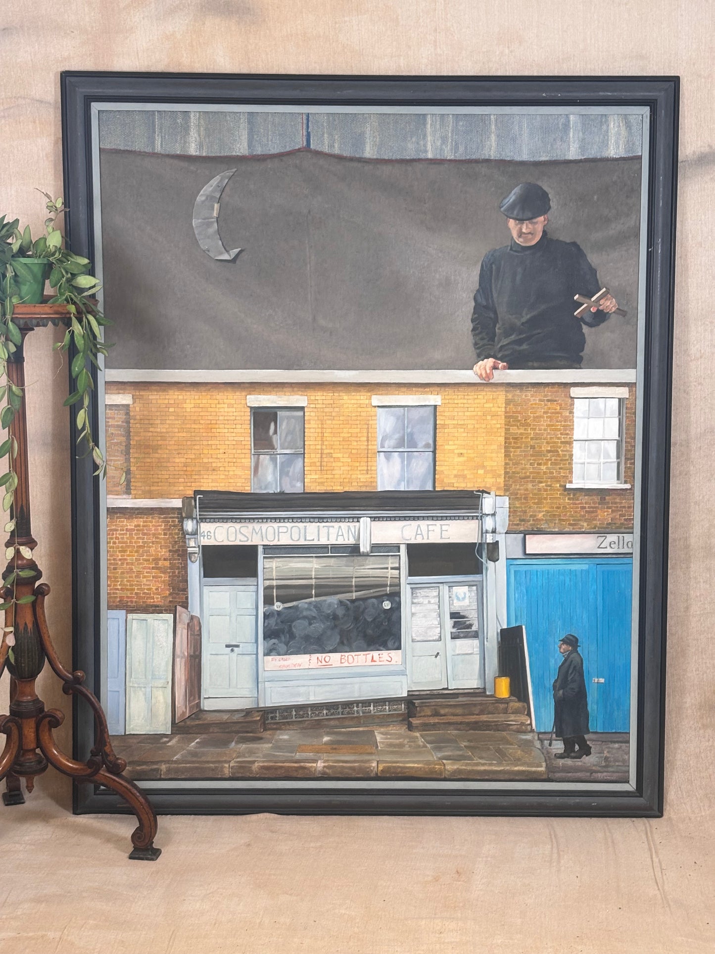 A framed art print depicting a street scene with a brick building, a blue door, and a person in black clothing, displayed against a beige wall next to a decorative wooden stand.