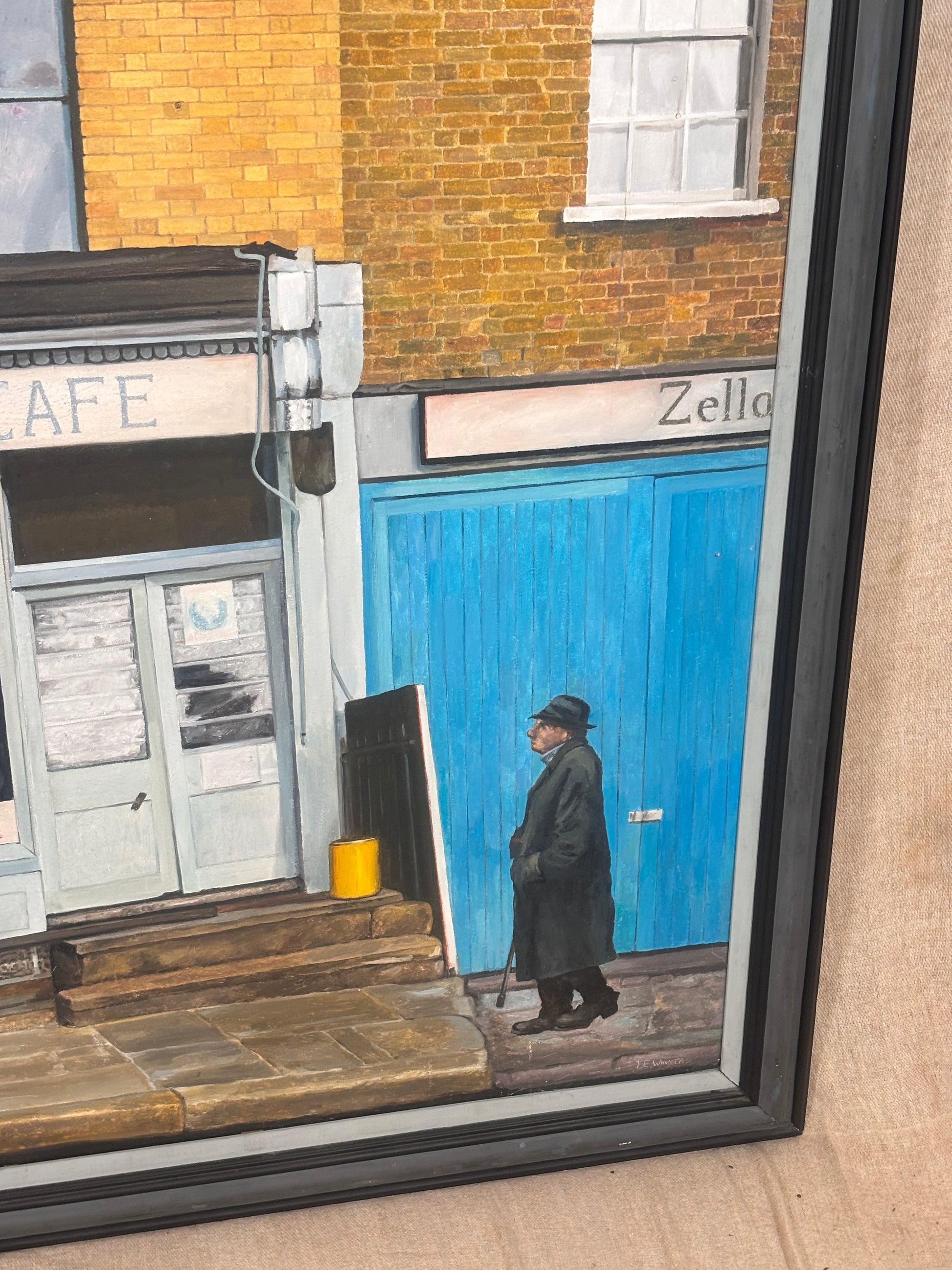 A framed painting depicting a street scene with a man in a long coat and hat walking in front of a bright blue shop door and a brick building.