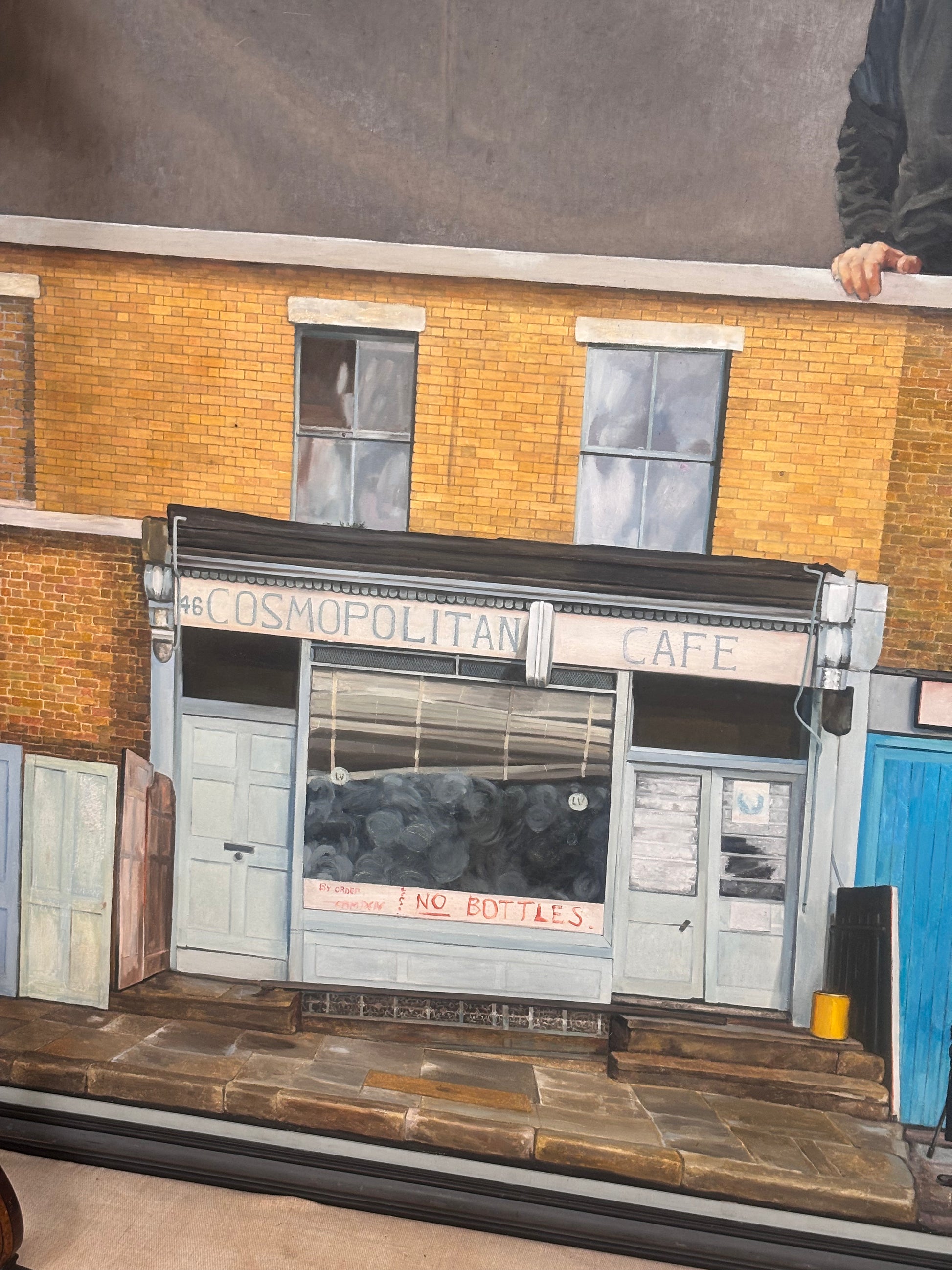 A painted brick building facade with a storefront reading 'The COSMOPOLITAN CAFE' above a window and doors, shown as artwork or a print. A small portion of a person's arm and hand is visible at the top right edge.