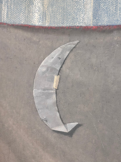 A close-up of a gray crescent moon applique or patch on a textured dark gray surface, with a blue and white patterned fabric strip and a thin red line visible at the top.