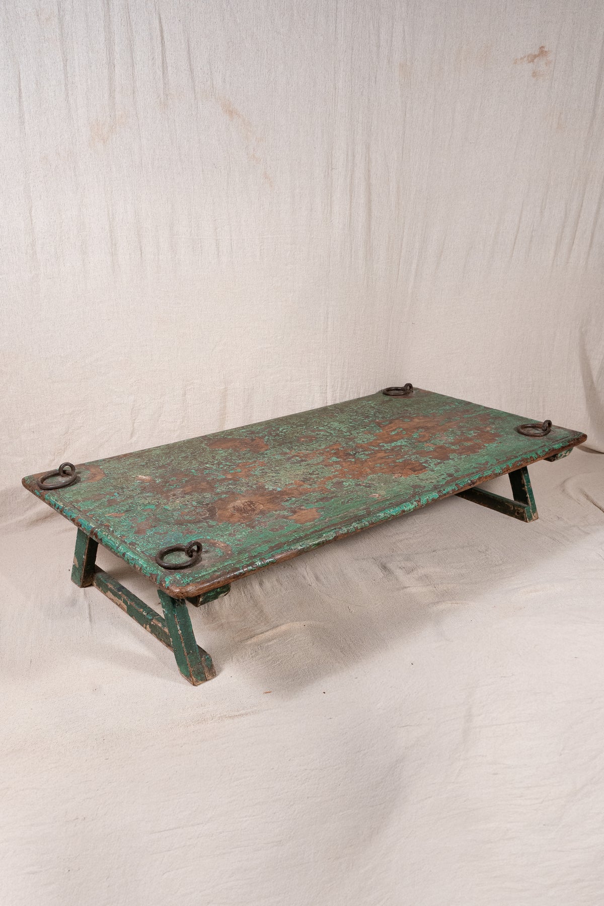 Low wooden bajot table with distressed green paint, angled legs and metal corner rings, possibly repurposed from an old gate or door.