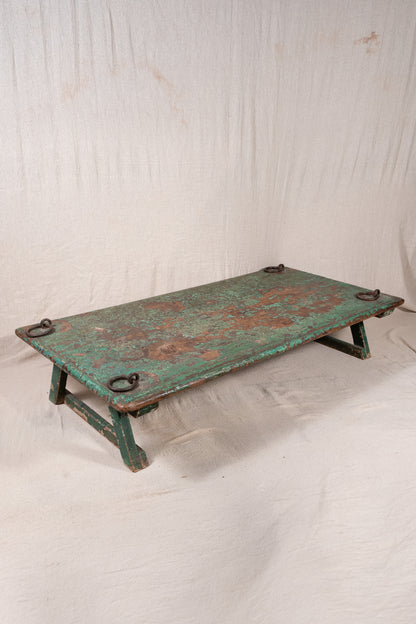 Low wooden bajot table with distressed green paint, angled legs and metal corner rings, possibly repurposed from an old gate or door.