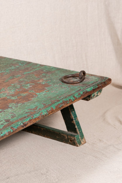 Low wooden bajot table with distressed green paint, angled legs and metal corner rings, possibly repurposed from an old gate or door.