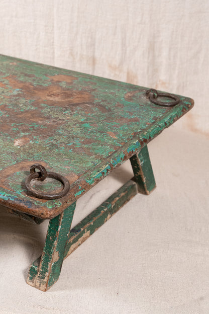 Low wooden bajot table with distressed green paint, angled legs and metal corner rings, possibly repurposed from an old gate or door.
