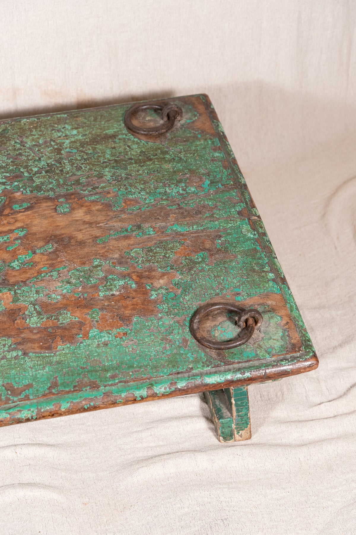 Low wooden bajot table with distressed green paint, angled legs and metal corner rings, possibly repurposed from an old gate or door.