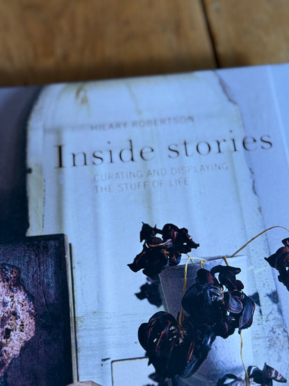 A close-up of a book titled 'Inside stories' by Heavy Robertson, featuring a light blue-gray cover with dark dried flowers and a rustic wooden surface in the background.