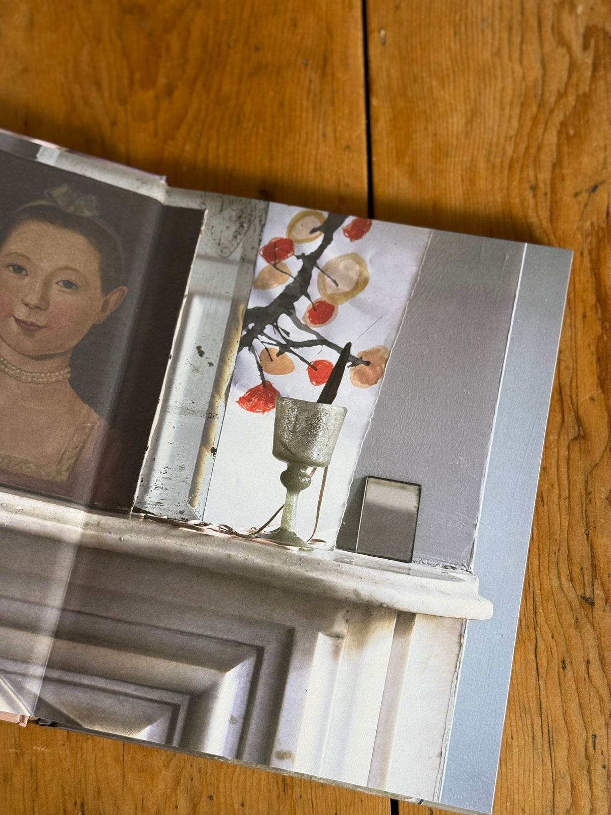 An open art book or magazine lying on a wooden floor, showing a printed still life scene with a painted portrait on one page and a still life floral/vase scene on the other.