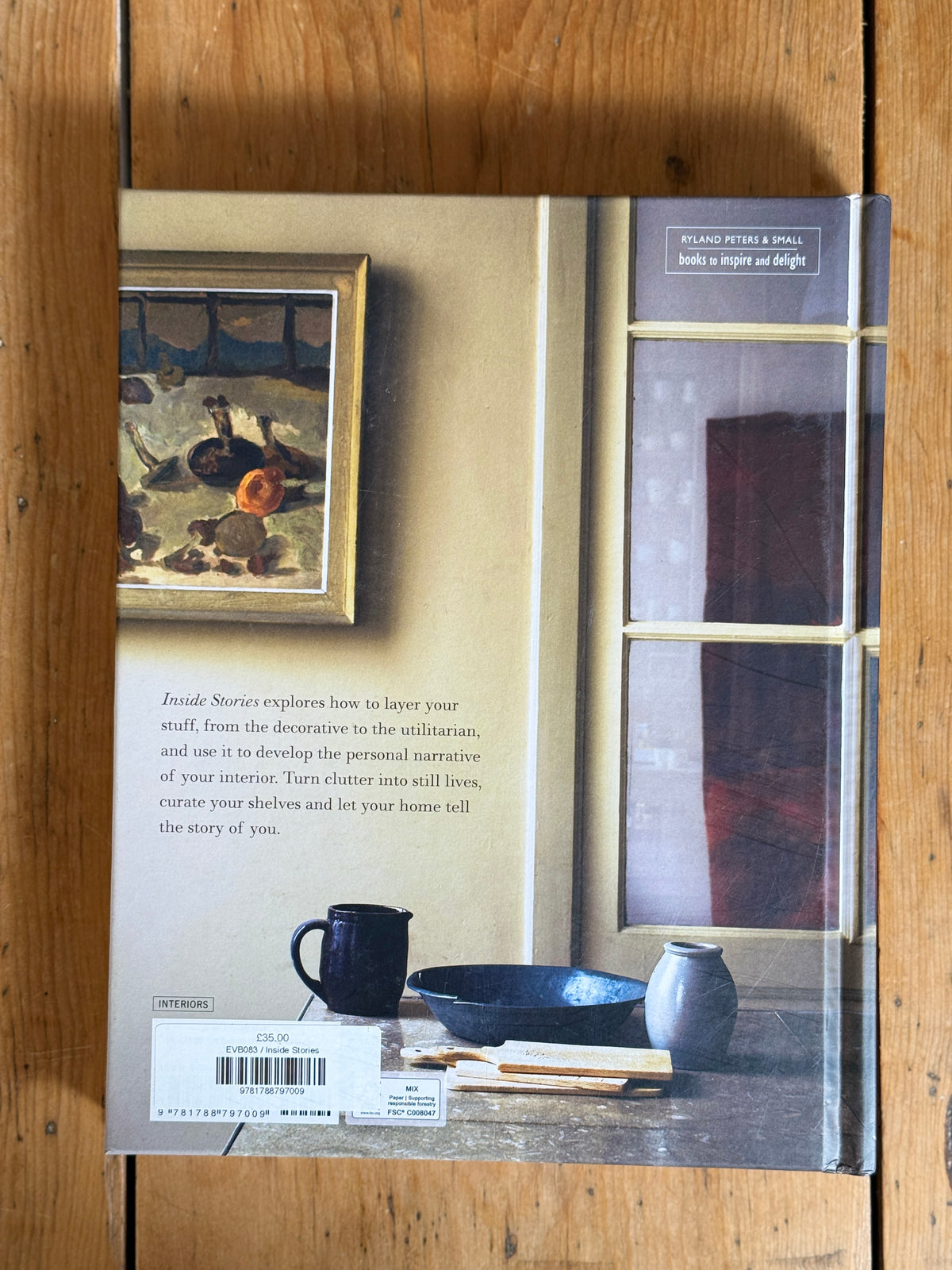 The back cover of a book titled 'Rustic Interiors: Building a Personal Narrative of Your Interior' by Richard Nelson & Sally, shown flat on a wooden surface. The cover features a warm beige background, a framed still life painting, a window view, and interior decor items like a mug, bowl, and vase.