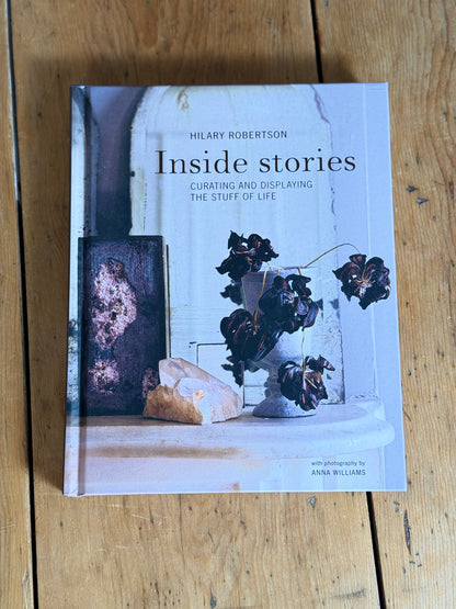 The book 'Inside stories: Curating and displaying the stuff of life' by Hilary Robertson, featuring a light blue-gray cover with a still life photograph of dried flowers and a rock, placed on a rustic wooden table.