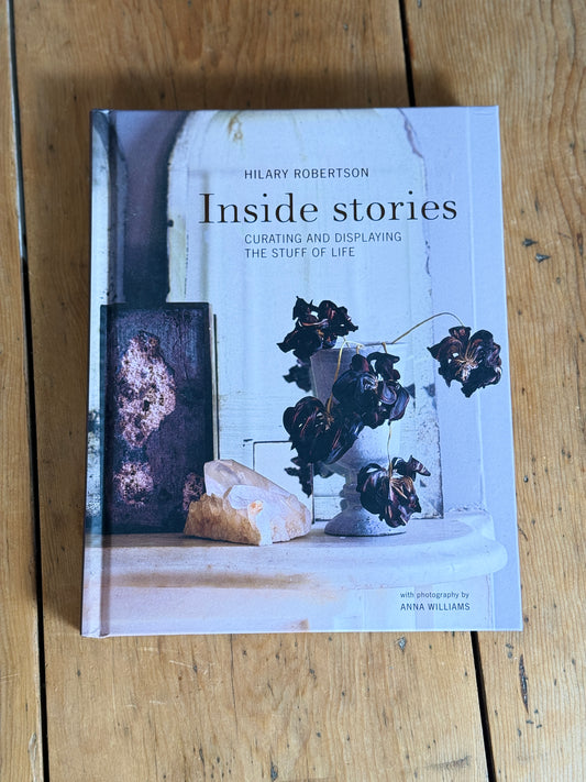 The book 'Inside stories: Curating and displaying the stuff of life' by Hilary Robertson, featuring a light blue-gray cover with a still life photograph of dried flowers and a rock, placed on a rustic wooden table.