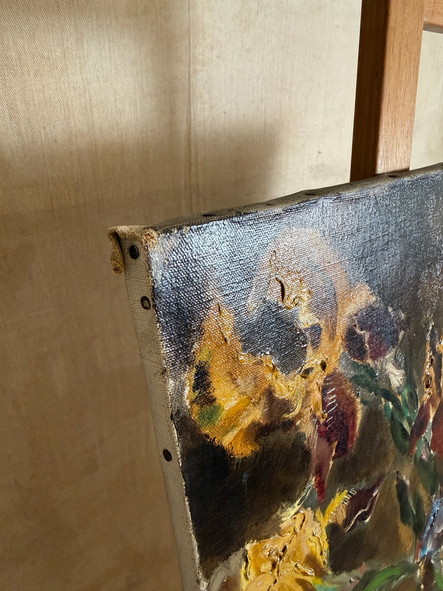 A close-up detail of an oil painting on canvas, showing thick impasto brushstrokes in dark blue, gold, brown, and green, mounted on a wooden easel against a beige wall.