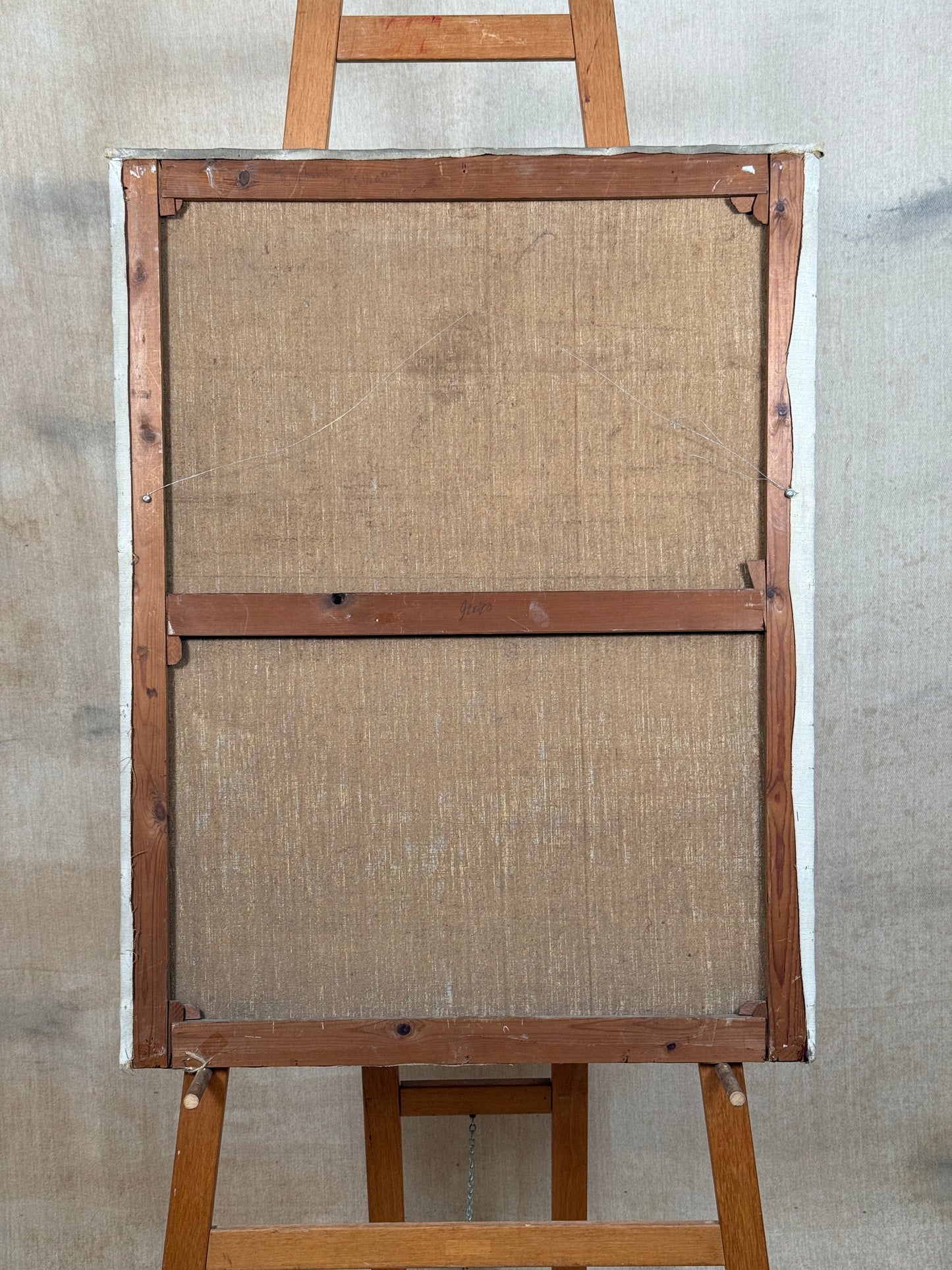The back side of a framed canvas print on a wooden easel, showing the canvas wrapped in white paper with a brown wooden frame and stretcher bars against a textured gray wall.