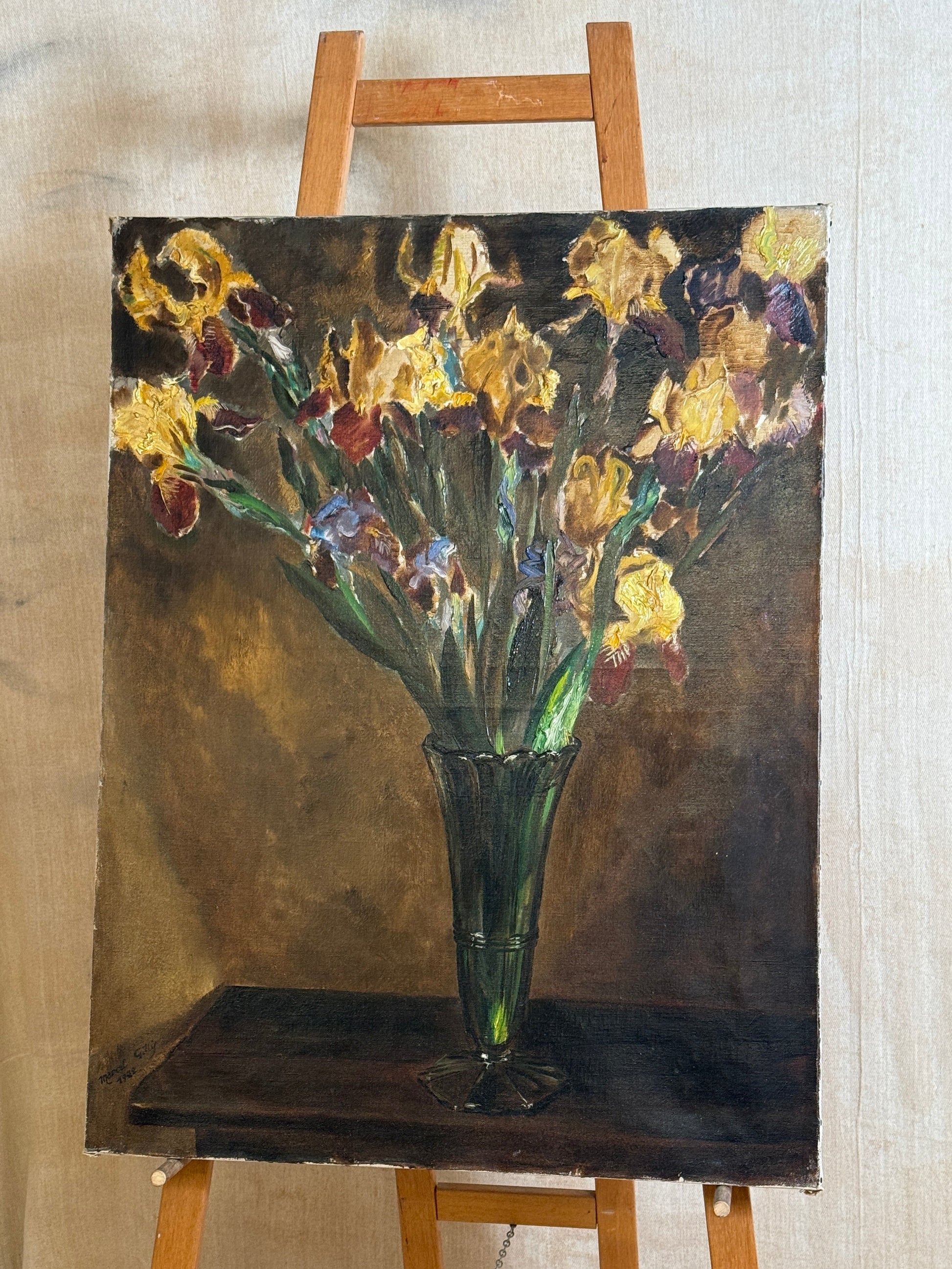 A canvas painting of yellow and purple irises in a dark green glass vase, displayed on a wooden easel against a light beige fabric backdrop.