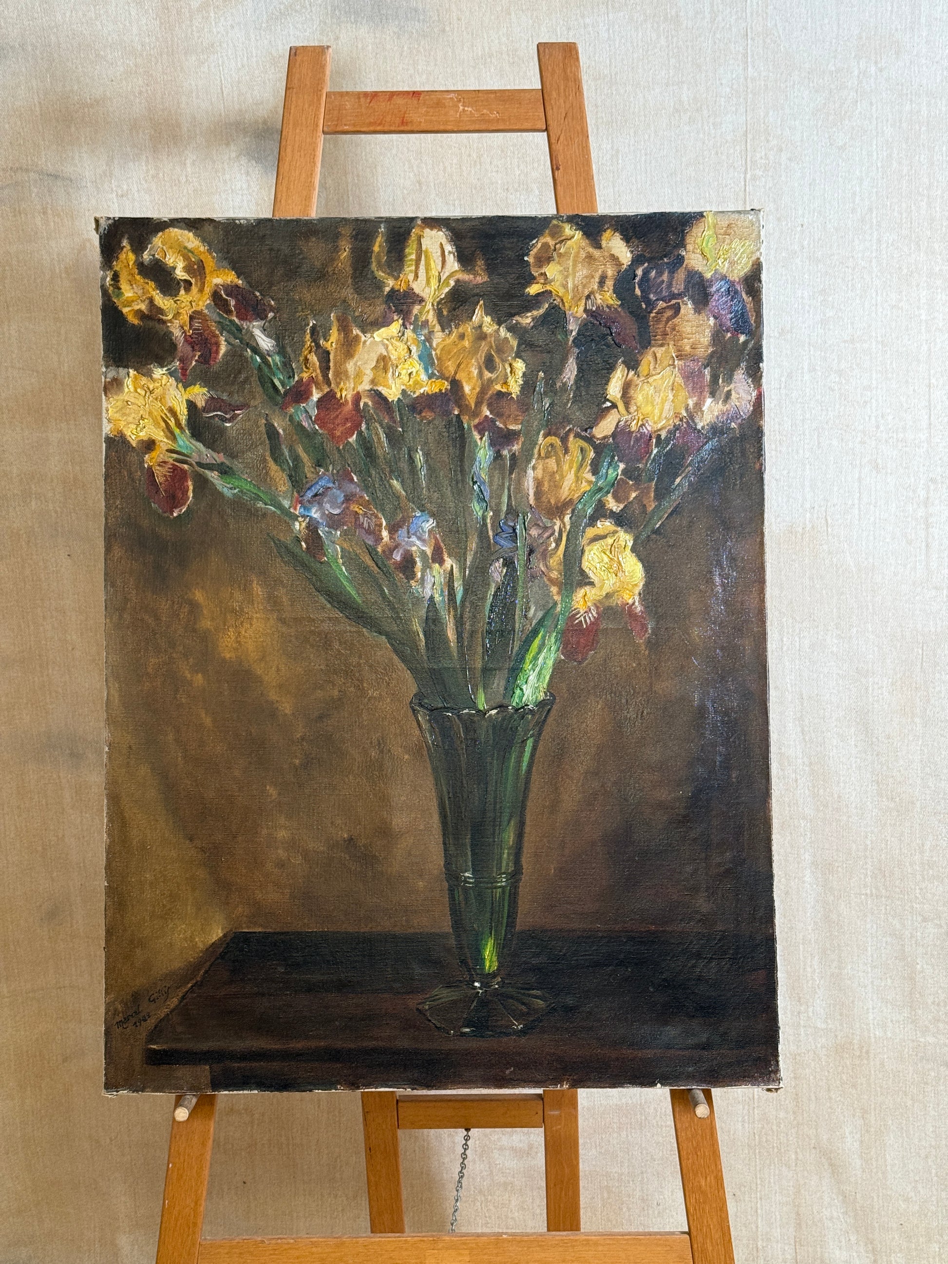 A vertical oil painting of yellow and purple irises in a green glass vase, displayed on a wooden easel against a neutral beige wall.