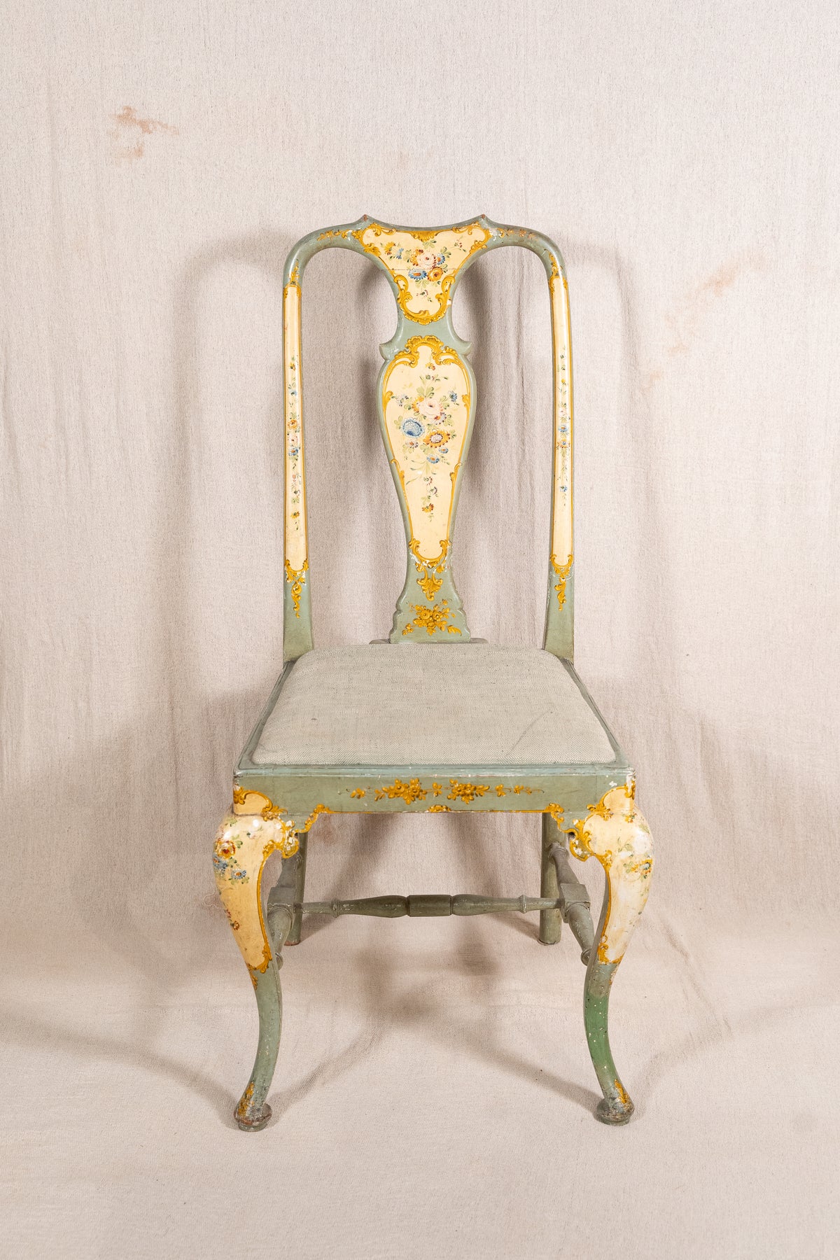 An antique-style metal dining chair with a distressed sage green and cream paint finish, featuring gold leaf accents and floral detailing on the backrest and legs, set against a neutral fabric backdrop.