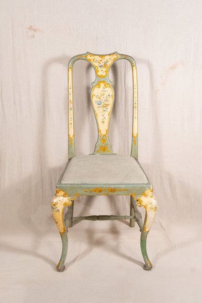 An antique-style metal dining chair with a distressed sage green and cream paint finish, featuring gold leaf accents and floral detailing on the backrest and legs, set against a neutral fabric backdrop.