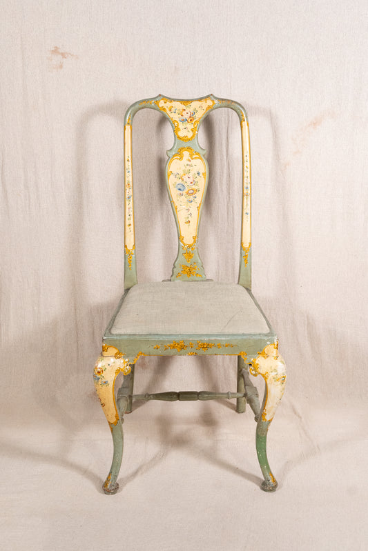 An antique-style metal dining chair with a distressed sage green and cream paint finish, featuring gold leaf accents and floral detailing on the backrest and legs, set against a neutral fabric backdrop.