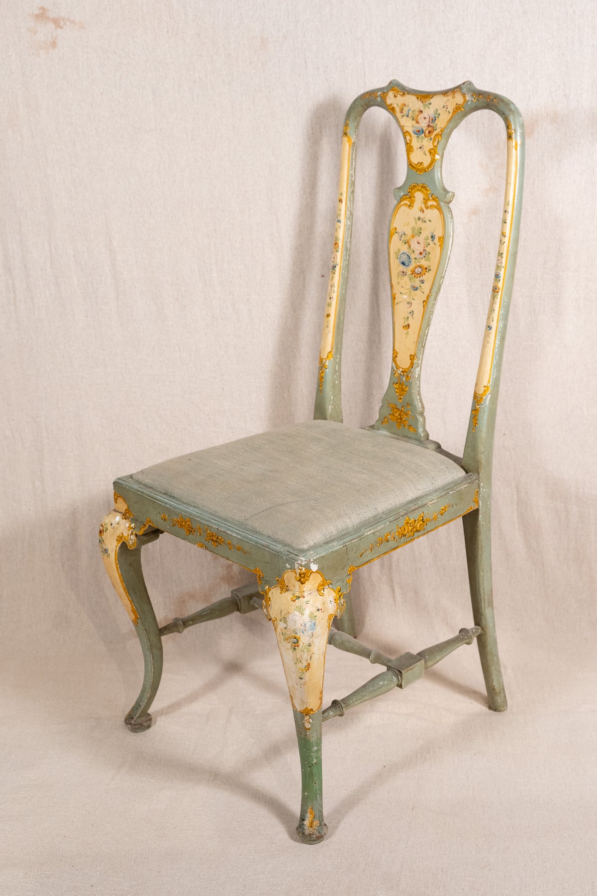 An antique French-style side chair with a distressed sage green and cream painted finish, featuring gold leaf detailing and a padded seat cushion, photographed against a neutral fabric backdrop.