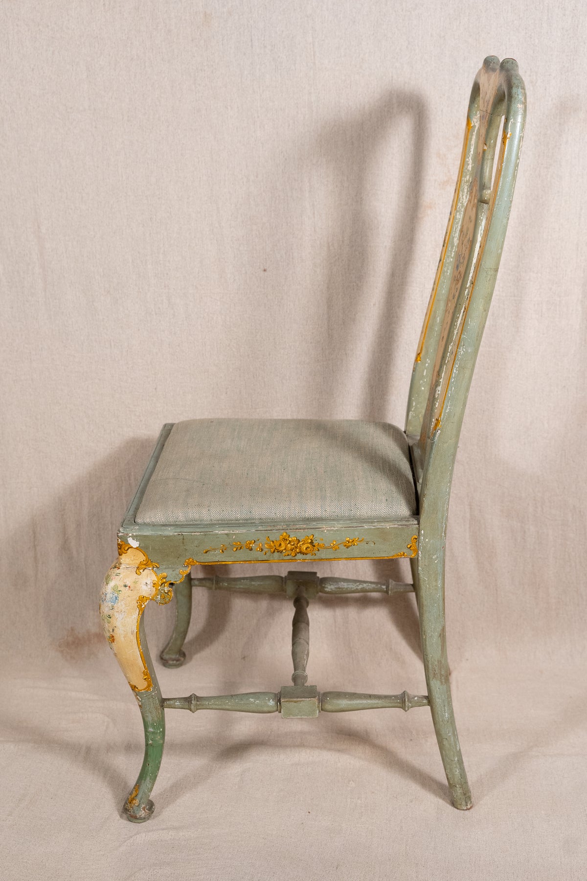 A distressed vintage wooden dining chair with a light sage green painted frame and seat cushion, showing significant yellow and white peeling paint on the legs and apron, set against a neutral beige fabric backdrop.