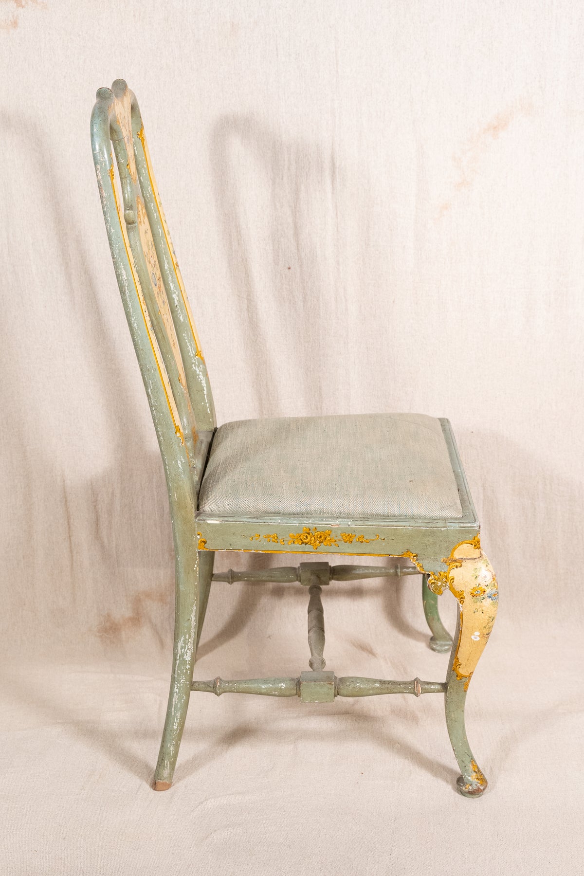 A distressed vintage wooden dining chair with a light sage green painted finish and peeling yellow-gold paint, featuring a padded seat cushion, photographed against a neutral off-white fabric backdrop.